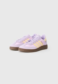 Pora šviesiai violetinių Adidas sportbačių su geltonomis juostelėmis ir rudais padais ant baltos fono, šiek tiek pasukta į kairę.