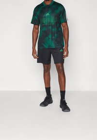 Zelené tričko Adidas s vzorovaným designem a síťovinou, spárováno s černými šortkami. Obsahuje logo a akcentní barvy. Nosit s černými sportovními botami.