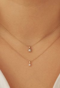 Two layered delicate gold necklaces with small round diamond pendants on a person's neck against light skin and white clothing.