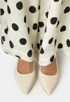 Pies con zapatos planos beige de punta afilada bajo una tela blanca con grandes lunares negros, sobre una superficie clara.