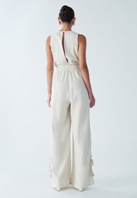 Svetlo bež kombinezon brez rokavov z odprtino na zadnji strani, nabranim pasom in širokimi hlačnicami s trakovi na robu.