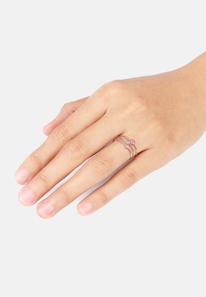Bague en or rose composée de trois anneaux, dont deux sont ornés de pierres précieuses roses et transparentes. Texture lisse et design épuré, portée sur une main.