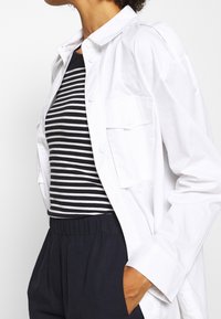 Camisa blanca de botones con mangas largas, que cuenta con bolsillos. Debajo, una blusa a rayas en blanco y negro. Combinada con pantalones oscuros de cintura elástica.