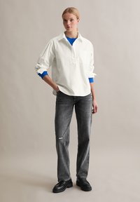 Witte oversized blouse met een borstzak, gedragen over een blauwe trui en gecombineerd met grijze distressed jeans en zwarte platform schoenen.