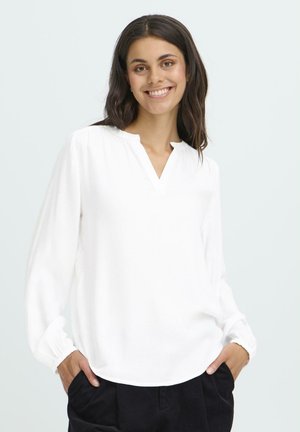 Smiling woman with dark hair wears a white long-sleeve blouse and black pants, standing with hands in pockets against a plain background.