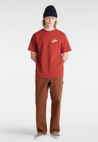 Camiseta de algodón roja con logo amarillo, de mangas cortas, con ribete blanco, combinada con pantalones marrones y zapatillas de cuadros. Gorro de lana azul marino.