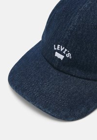 Gorra de béisbol de denim oscuro con el logotipo en texto blanco "LEVI'S". Presenta un ala curvada y una tela texturizada. Una visión general muestra los detalles de la costura.