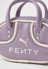 Geantă de piele texturată de culoare mov, mică, cu garnitură albă, cu două mânere și logo-uri albe Puma și Fenty pe față.
