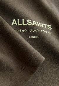 Camiseta de algodón marrón con el logo de “ALLSAINTS” en verde claro, texto en japonés y “LONDRES”. Primer plano de la textura y el detalle de la costura.