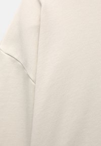 Gros plan d'un vêtement en tissu beige montrant un motif tricoté texturé et une couture cousue le long de la zone de l'épaule.