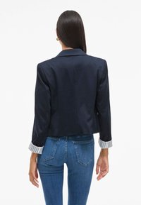 Tamsiai mėlynas švarkas su pritaikytu siluetu ir trijų ketvirčių rankovėmis. Audinys su tekstūra ir dryžuoti balti rankogaliai prideda detalių. Dėvima su mėlynais džinsais.