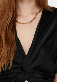 Collana in metallo dorato con maglie intrecciate, indossata su un top nero a scollo a V profondo, che mette in risalto una texture liscia e una finitura lucida.