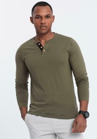 Man i olivgrön långärmad henley-skjorta med tre knappar, ljusgrå byxor och svart armbandsur, stående med en hand i fickan.