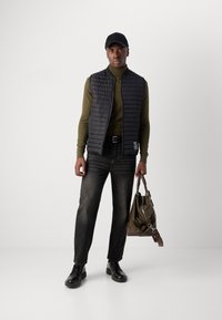Zwarte gewatteerde vest over een olijfkleurige coltrui, gecombineerd met donkere jeans en zwarte schoenen. Houdt een olijfgroene tas met een zilveren schouderband.