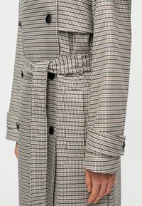 Μπεζ και μαύρο παλτό με μοτίβο κυνόδοντα, διπλό πέτο, μέση με ζώνη και μεγάλα κουμπιά· διαθέτει δομημένους ώμους και μανίκια με μανσέτες.