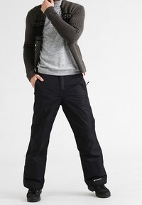 Mężczyzna w czarnych spodniach zimowych Columbia, szarej bluzie i brązowym kardiganie, stoi z jedną ręką w kieszeni, a drugą blisko twarzy.