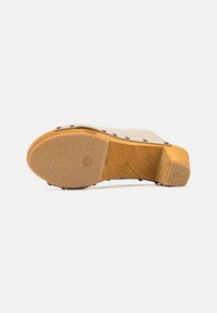 Sandale à talon en bois beige avec une semelle en caoutchouc texturée, dotée de clous en métal le long du bord supérieur et d'un talon étroit et incliné.