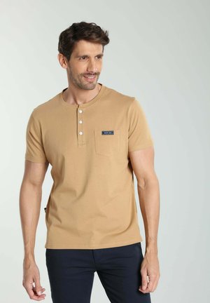 Homme portant un t-shirt henley beige à manches courtes avec une poche poitrine et un pantalon foncé, regardant sur le côté sur un fond uni.