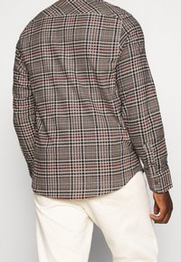 Hombre con una camisa de cuadros de manga larga en colores rojo, negro y beige, combinada con pantalones de color blanco roto, visto desde la espalda.