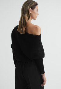 Zwarte geribbelde off-the-shoulder top met lange mouwen, gecombineerd met zwarte broeken. Gladde textuur en een ontspannen pasvorm, die een minimalistisch ontwerp laat zien.