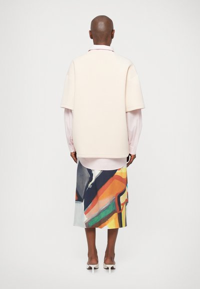 Kremna oversized majica s kratkimi rokavi, ki je prelečena preko svetlo rožnate srajce, v kombinaciji z midi krilom v barvnem abstraktnem vzorcu. Gladka tekstura tkanine.