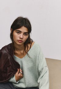 Pulover oversized de culoare verde deschis, cu decolteu adânc, purtat cu o jachetă din piele cărămizie aruncată pe umăr, evidențiind contrastul de textură.
