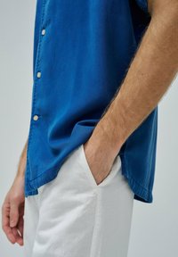 Camisa azul de manga corta hecha de tejido suave, con un cierre de botones en el frente y un corte relajado, combinada con pantalones de lino de color claro.