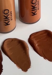 Three bottles of KIKO Milano foundation in varying shades of beige and brown, with swatches of foundation applied on a white surface.