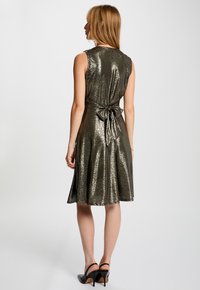 Knielange mouwloze jurk van metallic olijfgroen stof, met een strikband om de taille en een A-lijn rok met een subtiele textuur en glans.