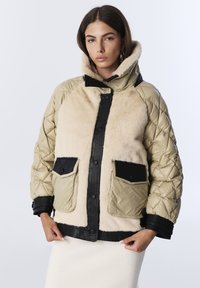 Femme portant une veste matelassée beige et noire avec de grandes poches avant en fausse fourrure, se tenant devant un fond clair.