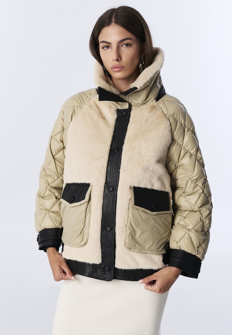 Femme portant une veste matelassée beige et noire avec de grandes poches avant en fausse fourrure, se tenant devant un fond clair.