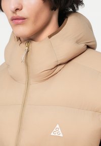 Chaqueta de plumas acolchada beige con cuello alto, cierre frontal y un pequeño logotipo blanco. Textura suave con un brillo sutil. Diseño ligero.