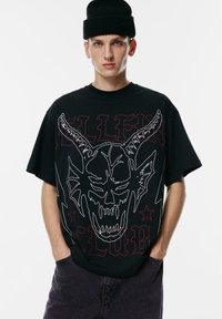 Mladic u crnoj kapici i prevelikoj crnoj majici s bijelim konturama rogate lubanje i crvenim gotičkim tekstom, pozira s rukama u džepovima tamnih hlača.