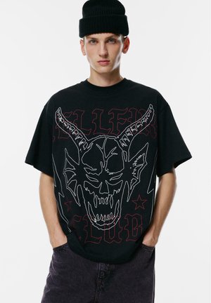 Mladý muž v čiernej čiapke a prehnanom čiernom tričku s bielym kontúrovaným lebkovým motívom a červeným gotickým textom, ktorý pózuje s rukami v tmavých nohaviciach.