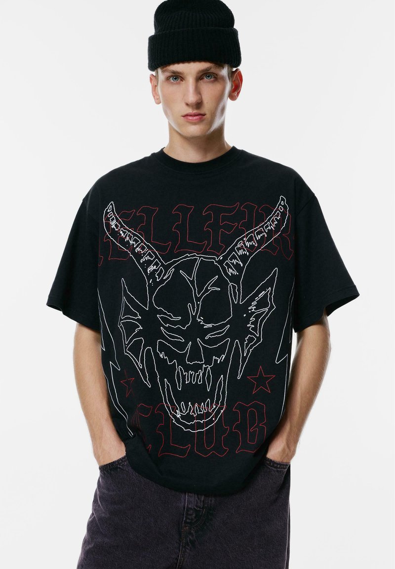 Mladic u crnoj kapici i prevelikoj crnoj majici s bijelim konturama rogate lubanje i crvenim gotičkim tekstom, pozira s rukama u džepovima tamnih hlača.