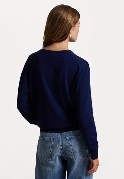 Woman with long brown hair wearing a navy blue sweater and light blue jeans, shown from behind against a plain white background.