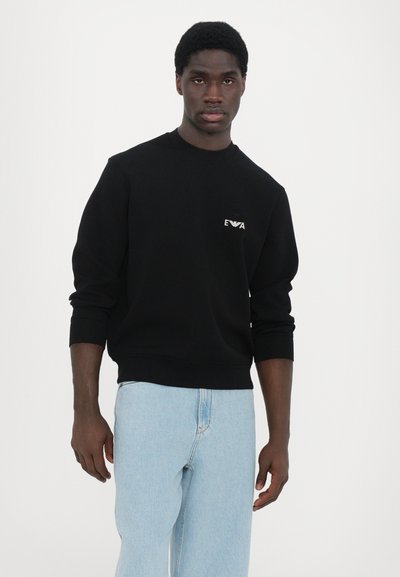 Sweatshirt noir à manches longues, col rond et poignets côtelés. Présente un petit logo blanc sur la poitrine. Porté avec un jean bleu clair.