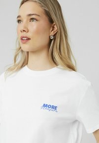 T-shirt en coton blanc avec un texte graphique bleu "PLUS de Plaisir" sur la poitrine. Manches courtes et col rond classique.