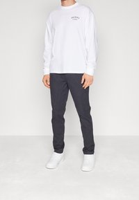 Bílé tričko s dlouhým rukávem s malým černým logem, doplněné tmavě šedými kalhotami a bílými teniskami, s přiléhavým střihem.