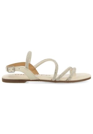 Sandale plate beige avec des fines lanières scintillantes et une boucle ajustable à la cheville sur une semelle marron, vue de côté sur fond blanc.