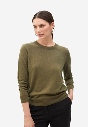 Suéter verde oliva con cuello redondo, puños acanalados y una textura suave, combinado con pantalones oscuros. El modelo está de pie ligeramente de lado.