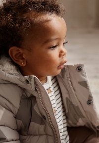 Veste matelassée gris clair avec motif géométrique, doublée d'un matériau doux de couleur crème, portée par-dessus une chemise à rayures blanches et bleu marine avec des boutons en bois.