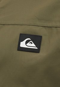 Μαύρο ορθογώνιο έμπλαστρο με λευκό κύμα και λογότυπο βουνού ραμμένα σε ελιάς χρώματος ύφασμα.