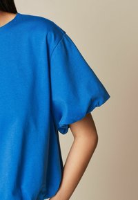 Modrý top s krátkým rukávem a volným střihem. Měkká textura, nafouknuté rukávy a kulatý výstřih. Jednoduchý design bez dalších vzorů nebo akcentů.