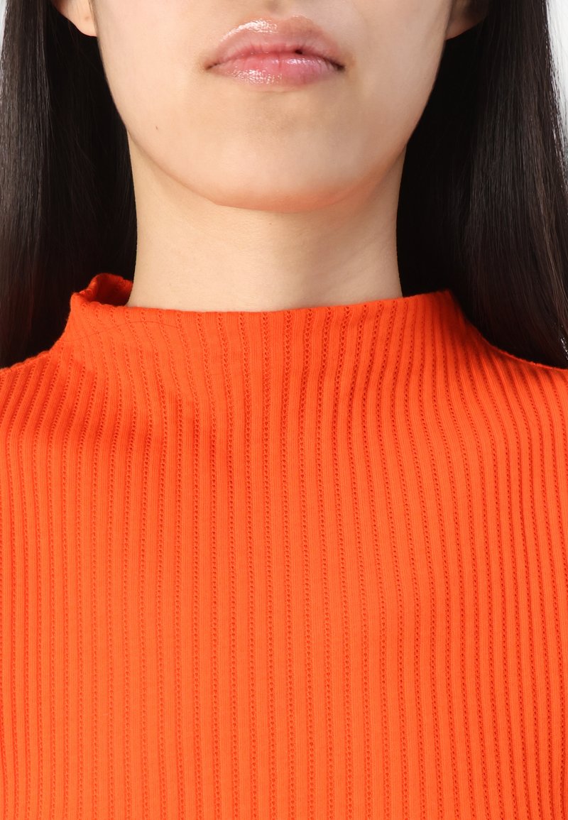 Femme aux longs cheveux noirs portant un pull côtelé orange à col montant, montrant ses lèvres et le bas de son visage ainsi que son cou.