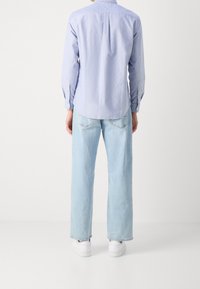 Persoon die met de rug naar de camera staat, gekleed in een lichtblauw overhemd met knopen, lichtgewassen spijkerbroek en witte sneakers, tegen een effen witte achtergrond.