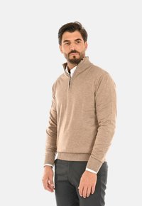 Maglione beige lavorato a maglia con collo zip e polsini e orlo a coste. Indossato sopra una camicia bianca, abbinato a pantaloni scuri.