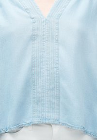 Ενδύμα σε ανοιχτό γαλάζιο με V-λαιμόκοψη, με κάθετες ραφές στο κέντρο, φορεμένο πάνω από λευκό παντελόνι.