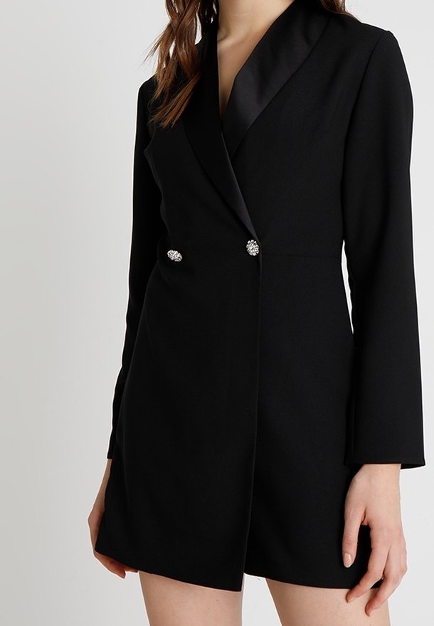 Mujer con un vestido tipo blazer negro ajustado, con solapas de satén y dos botones decorativos de pedrería en la cintura.