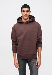 Νεαρός άνδρας φοράει ένα μοβ hoodie με μπροστινή τσέπη και γκρι τζιν, στέκεται μπροστά σε έναν απλό φωτεινό φόντο.